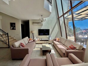 A modern living room with a large window, a flat-screen TV, and a staircase. - Villa Meltem Luxury Villa with Private Pool & Panoramic Sea Views – Kalkan (Antalya)