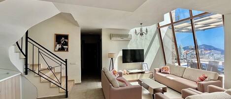 A spacious living area with a staircase, a flat-screen TV, and a large window offering a city view.