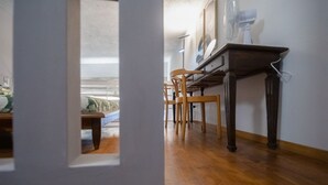 Una habitación con piso de madera, una mesa de comedor con sillas, una cama con manta verde y un ventanal con vista a la playa.
