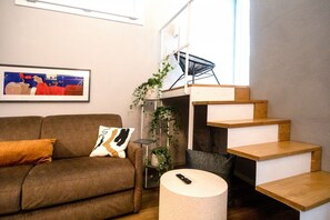Un salon avec un canapé marron, un tableau encadré au mur, une chaise noire, un escalier et une table ronde avec une télécommande.