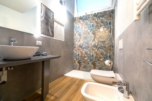 Une salle de bain moderne dotée d’un grand lavabo sur pied, d’un autre lavabo séparé, de toilettes et d’une douche avec un mur décoré d’un motif de feuilles.