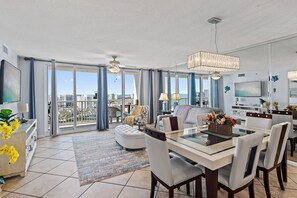 A spacious living area with a dining table, chairs, and a sofa. There is a large window with curtains, a ceiling fan, and a chandelier. - 🌴Walk to Beach | Gulf Views | Pelican Terrace 1404🌴 (Destin)