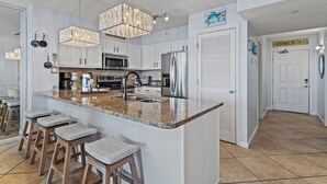 A modern kitchen with white cabinets, a granite countertop, and stainless steel appliances. There are bar stools and a chandelier. - 🌴Walk to Beach | Gulf Views | Pelican Terrace 1404🌴 (Destin)