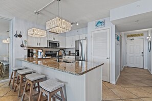 A modern kitchen with white cabinets, a granite countertop, and stainless steel appliances. There are bar stools and a chandelier. - 🌴Walk to Beach | Gulf Views | Pelican Terrace 1404🌴 (Destin)
