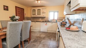 A kitchen with white cabinets, a dining table with chairs, and a countertop with a bread loaf and tomatoes. - Wheelhouse Cottage (Whitby)