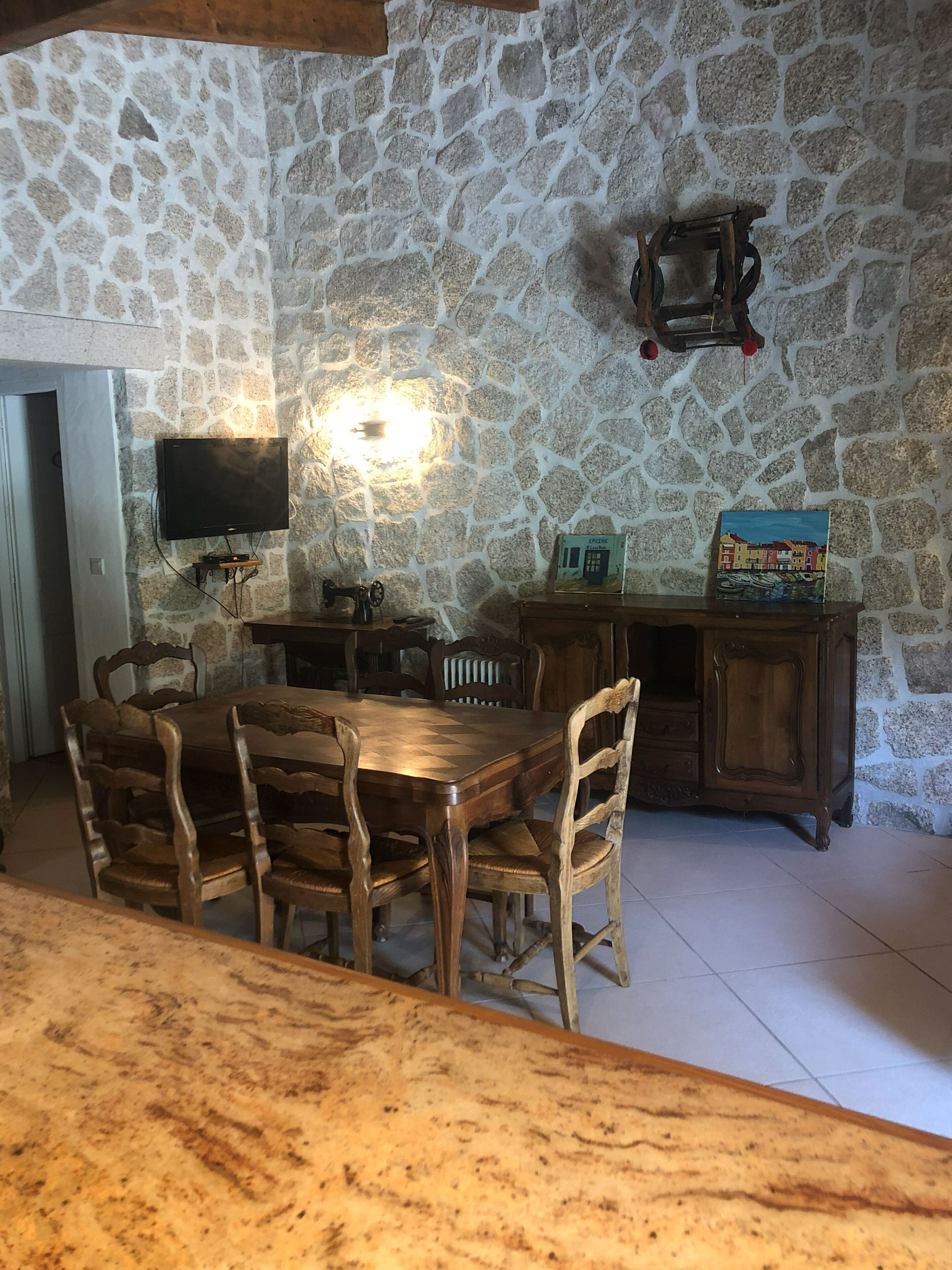 Una habitación con paredes de piedra, una mesa de comedor de madera y sillas, un televisor montado en la pared y un mueble con objetos decorativos.