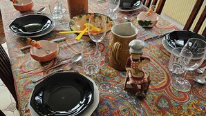 Une table à manger dressée avec des assiettes, des verres et une bouteille, entourée de chaises et recouverte d’une nappe à motifs.