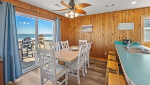 Een houten interieur met een eettafel, stoelen, een plafondventilator en uitzicht op de oceaan door een glazen deur.