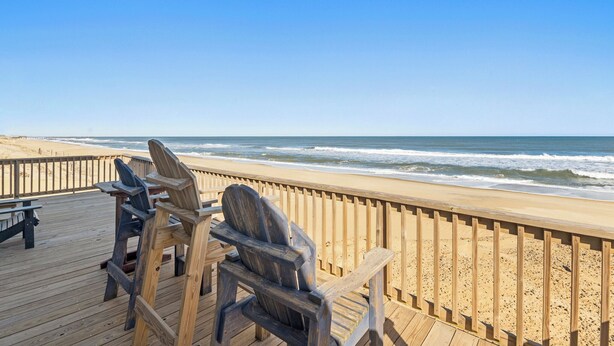 Een houten terras met Adirondack-stoelen met uitzicht op een zandstrand en de oceaan.