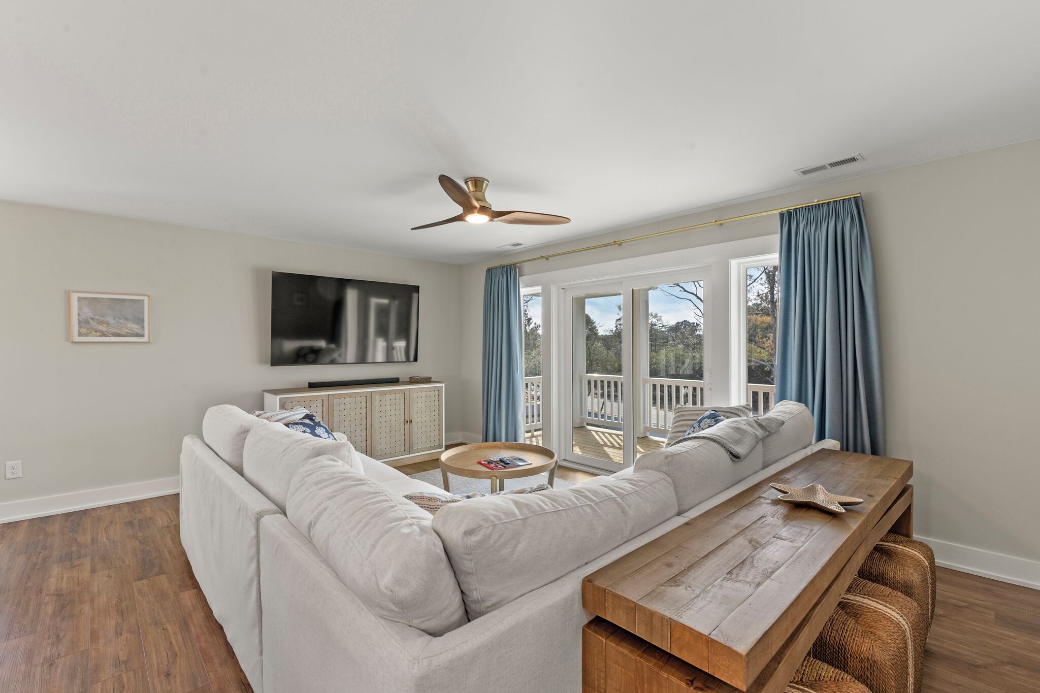 A living room with a sofa, a wooden coffee table, a ceiling fan, a TV, and a view through large windows.
