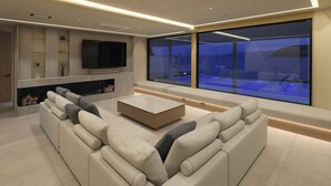 A modern living room with a large sectional sofa, a flat-screen TV, and a glass wall offering a view of the outdoors.