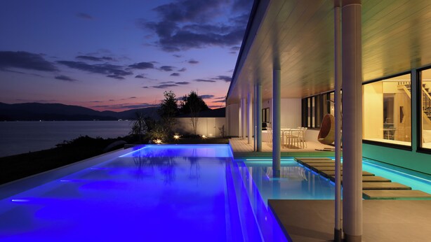 A modern house with an infinity pool, a view of the water, and a patio area with outdoor furniture.