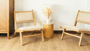 Ein Zimmer mit zwei Holzsesseln, einem kleinen runden Tisch mit einer Vase und einem Holzschrank mit Fischgrätenmuster.