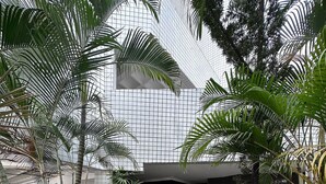 Uma passagem leva a uma entrada coberta, com paredes em azulejo e grades de metal, cercada por diversas plantas e árvores.
