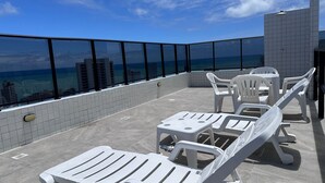 Terraço no telhado com cadeiras e mesas de plástico branco, piso ladrilhado e guarda-corpo de vidro com vista para a paisagem urbana e o mar.