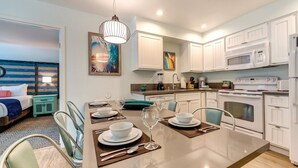 A modern kitchen with white cabinets, a dining area with a table set for four, and a bedroom with a bed and a nightstand.