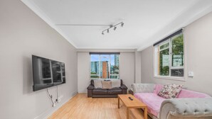 Sala de estar com televisão de tela plana na parede, sofá de couro marrom, sofá cinza com detalhes em botão e mesa de centro de madeira, além de janela com vista para edifícios altos.