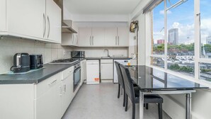 Uma cozinha compacta com armários brancos, bancada preta e um espaço de refeições com mesa e cadeiras. Uma janela oferece vista para a cidade.