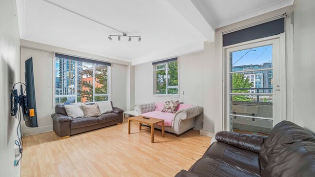 Uma sala de estar ampla com piso de madeira, dois sofás, uma mesa de centro e grandes janelas com vista para edifícios e árvores.