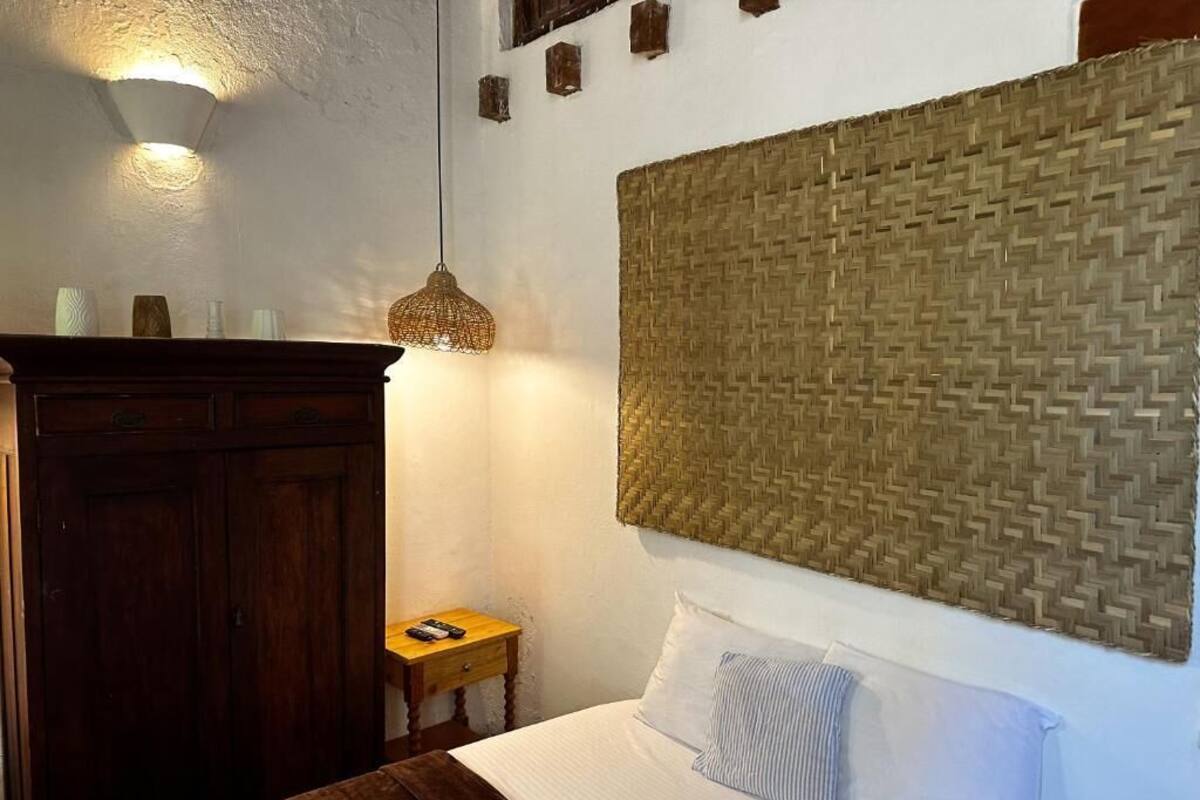 Un dormitorio con cama, armario de madera, mesita de noche, cabecero tejido y lámpara de pared.