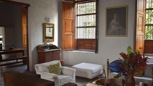 Ein Wohnzimmer mit einer Couch, einem Sessel und einem kleinen Tisch mit Dekoration. Ein Fenster mit Gitter und ein Bild an der Wand sind ebenfalls zu sehen.