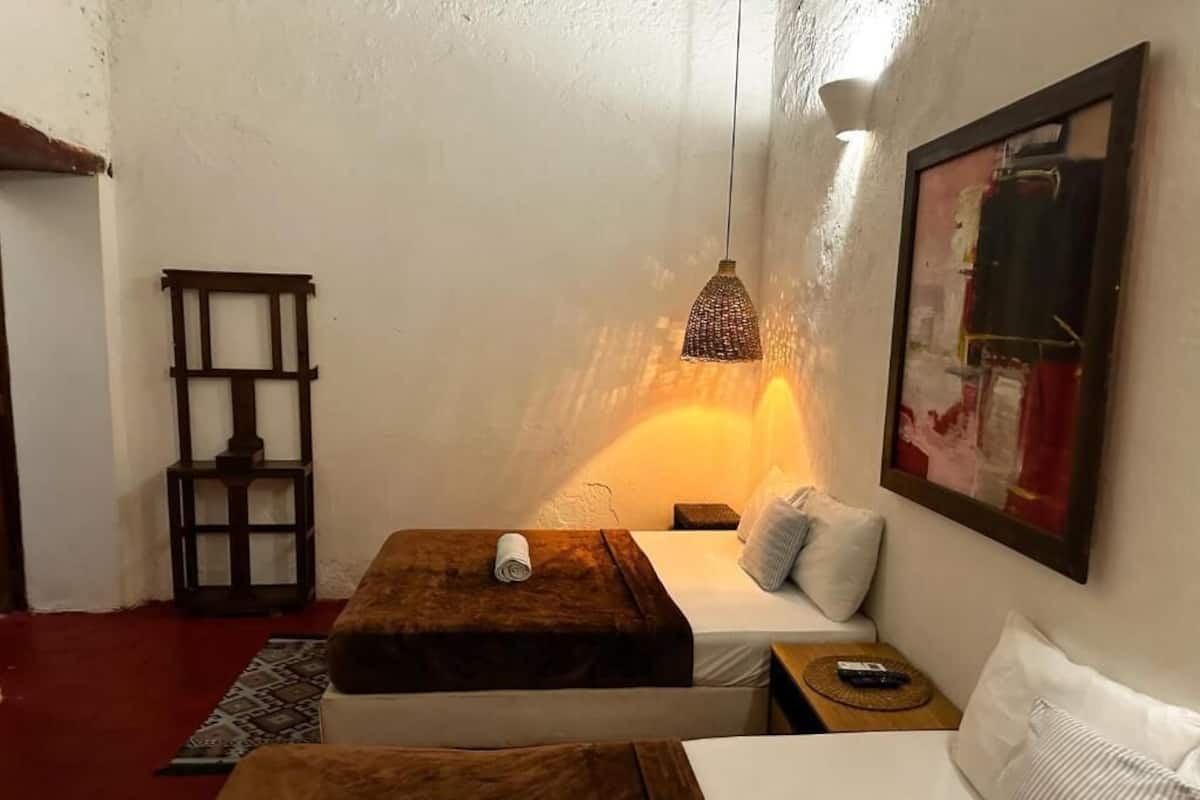Una habitación con dos camas, una lámpara colgante, un cuadro y una mesita con un control remoto.