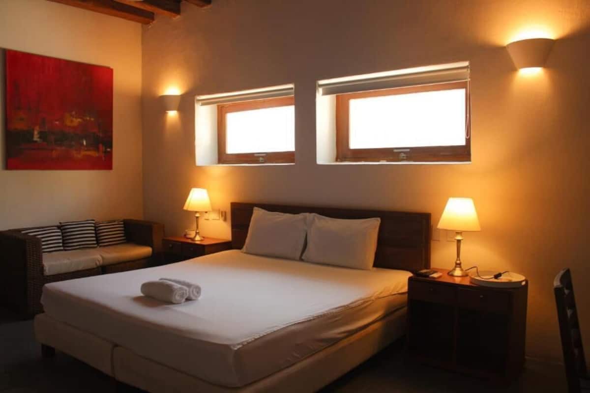 Una habitación de hotel con una cama grande, dos lámparas de noche, una mesita de noche, un sofá y dos ventanas con luces empotradas.