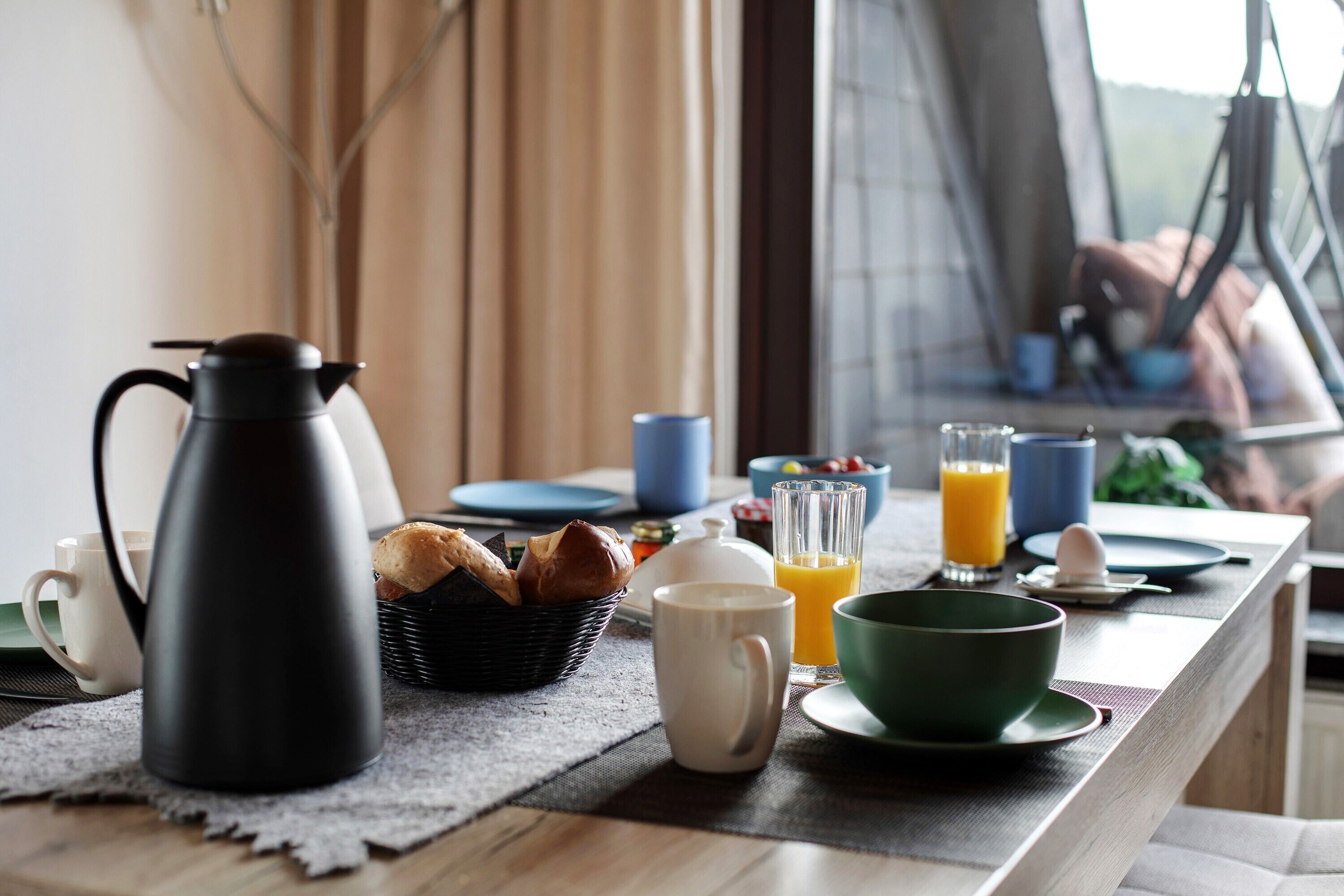 En morgenmadsopsætning med kaffekande, brød, en skål, en kop og et glas appelsinjuice på et bord.