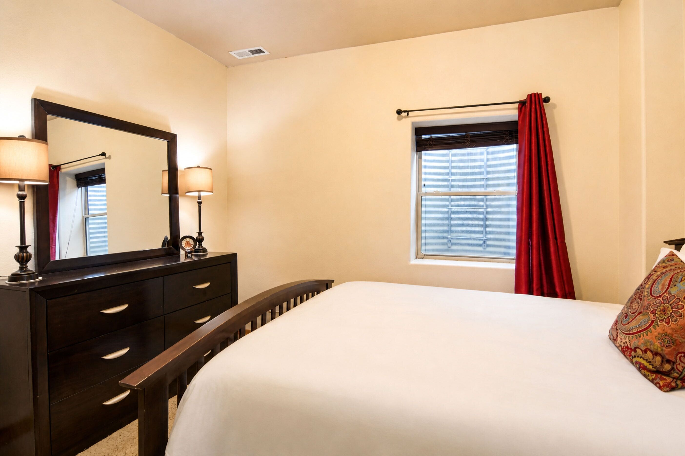 Een slaapkamer met een bed, een dressoir, een spiegel, een raam met jaloezieën en een rood gordijn.