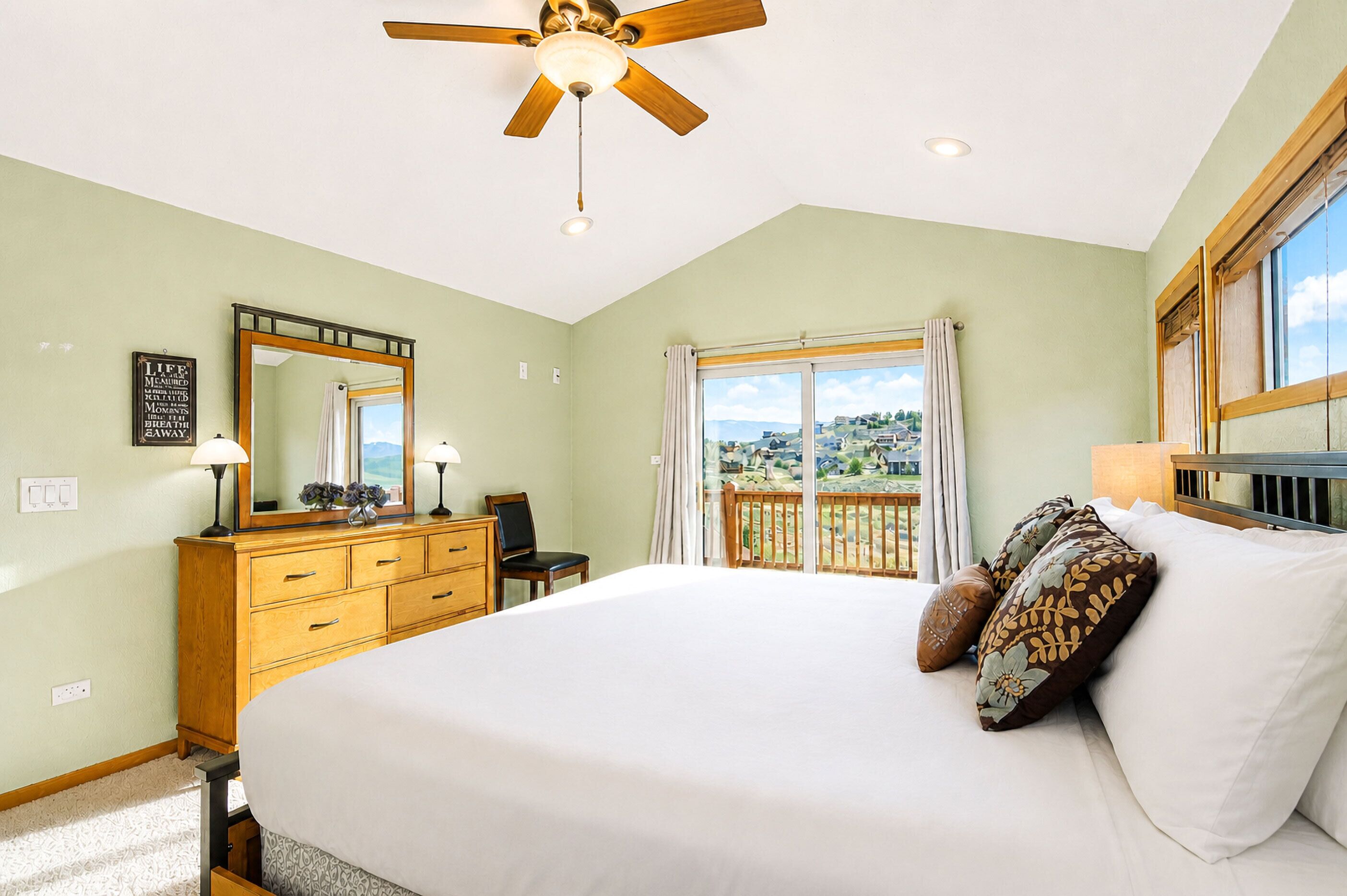 A bedroom with a bed, dresser, mirror, ceiling fan, and a view of the outside.