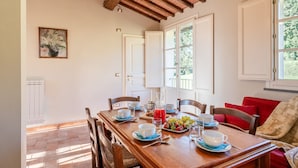 A dining area with a wooden table set for breakfast, white walls, and a view of greenery outside. - Romeo Apartment with Swimming Pool (Capannori)