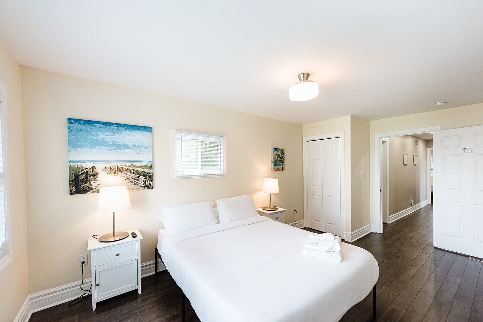 Une chambre d’hôtel avec un lit, des lampes de chevet, un tableau, une fenêtre avec des stores et une porte.