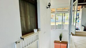 Una entrada limpia y bien iluminada, con una puerta blanca, un ventanal pequeño con cortina, un radiador, una planta en maceta y suelo de baldosas.