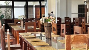 Uma longa mesa de jantar com cadeiras de madeira e jogos americanos trançados, em um ambiente com grandes janelas e um vaso de flores.
