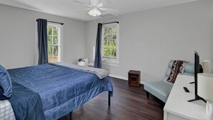 A bedroom with a bed, a desk, a chair, a ceiling fan, and two windows with curtains. - The City Cottage (West Columbia)