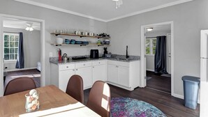 A modern kitchen with white cabinets, a granite countertop, and a ceiling fan. There is a dining area with a table and chairs, and a trash can is visible. - The City Cottage (West Columbia)