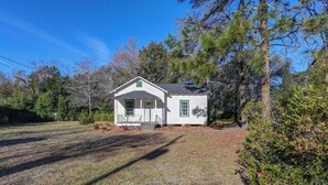 A white single-story house with a front porch, surrounded by trees and a grassy yard. - The City Cottage (West Columbia)