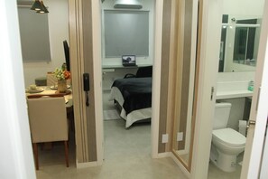 A hotel room with a bed, a desk with a computer, a small table with a vase, and a bathroom with a toilet and mirror. - Paradise in Lapa - Downtown Rio de Janeiro, Close to Everything + Wi-Fi (Rio de Janeiro)