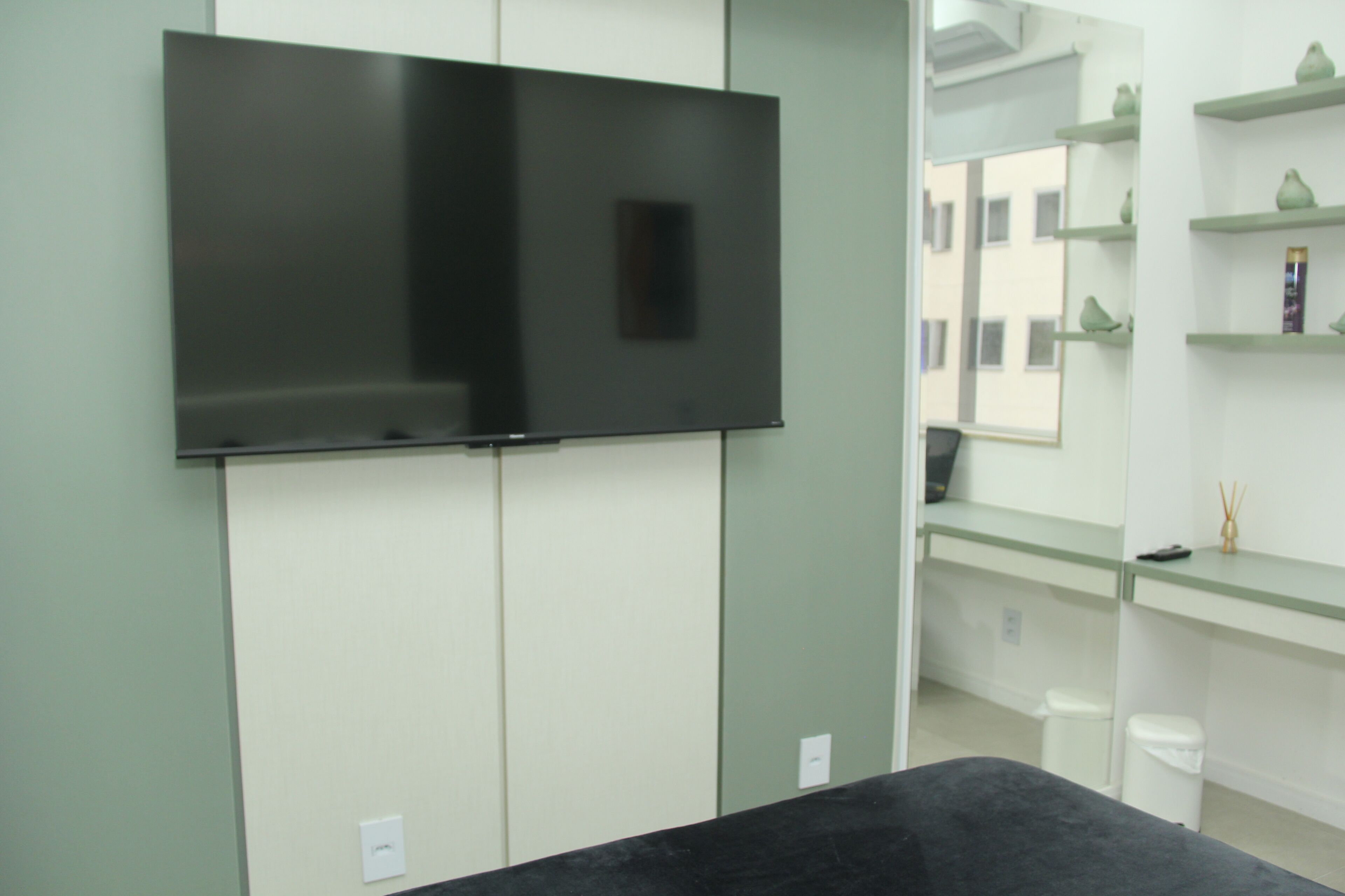 Quarto moderno com televisão de tela plana na parede, mesa preta e prateleira de vidro com objetos decorativos.