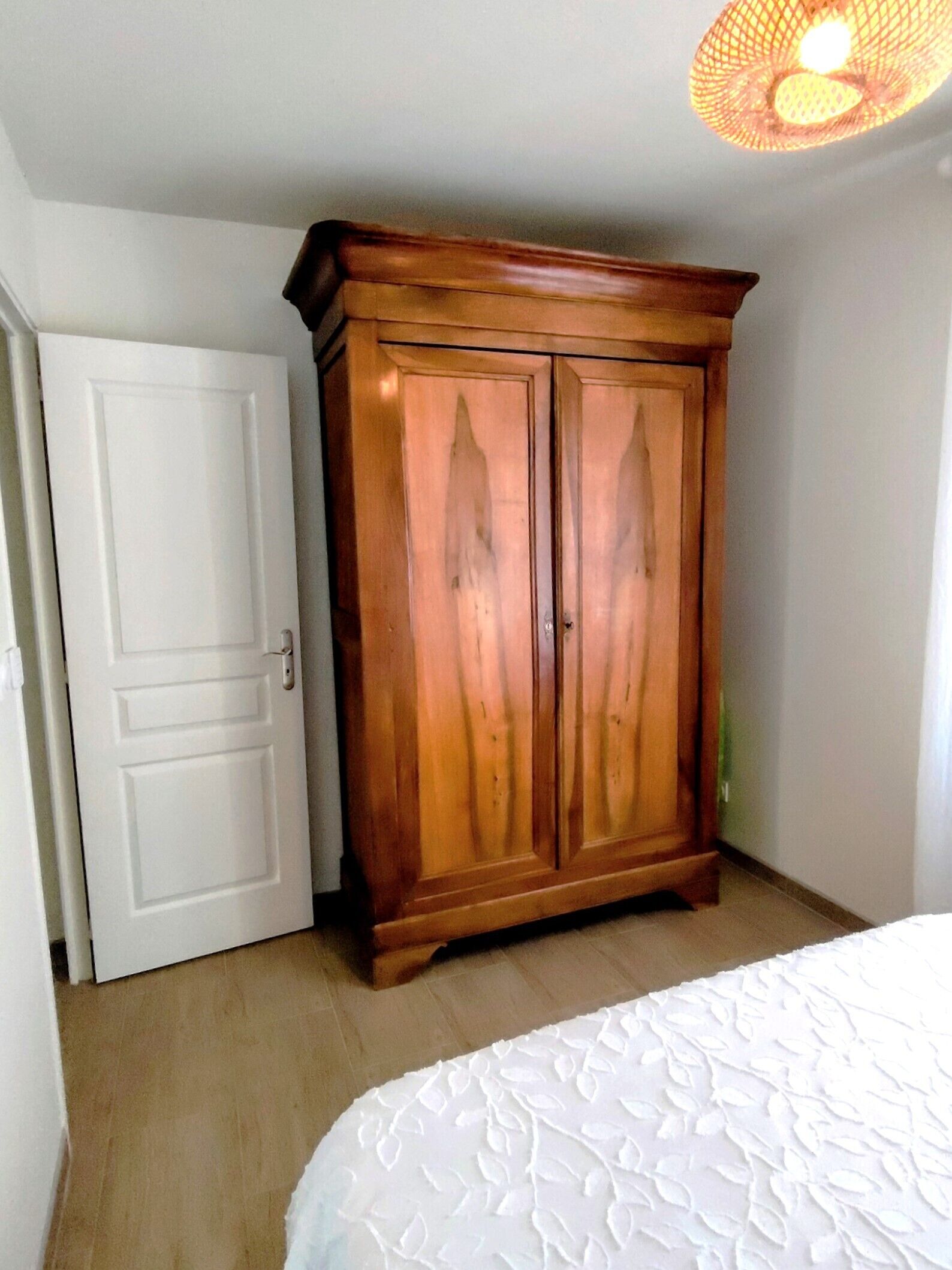Une chambre avec une armoire en bois, une porte blanche et un lit recouvert d’une couette blanche.