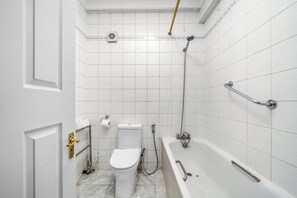 Een badkamer met witte betegelde wanden, een toilet, een badkuip met douchekop en een handgreep.