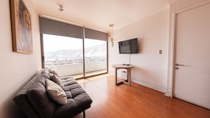 A modern living room with a sofa, a TV mounted on the wall, and a large window offering a city view.