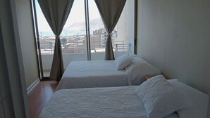 A hotel room with two beds, a view of the city and mountains through large windows, and a balcony with a railing.
