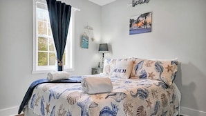 Een slaapkamer met een bed, voorzien van een dekbedovertrek met een strandthema, een raam met gordijnen, een nachtkastje met een lamp, en een muur met een bordje 'beach' en een lijst met een foto.