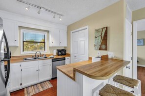 A modern kitchen with white cabinets, a wooden countertop, a built-in dishwasher, and a window with blinds. - Carolina Breeze - Walk to Carolina Beach (Carolina Beach)