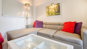 A modern living room with a grey sectional sofa, a glass coffee table, and a wall-mounted painting of a forest. - BELAIR (St. Julian's)