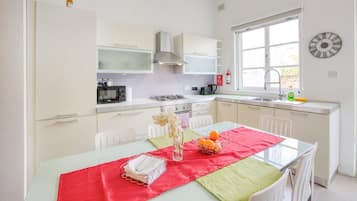 A modern kitchen with white cabinets, a stainless steel range hood, a microwave, and a gas stove. There is a window with a view of the outdoors, and a wall clock.