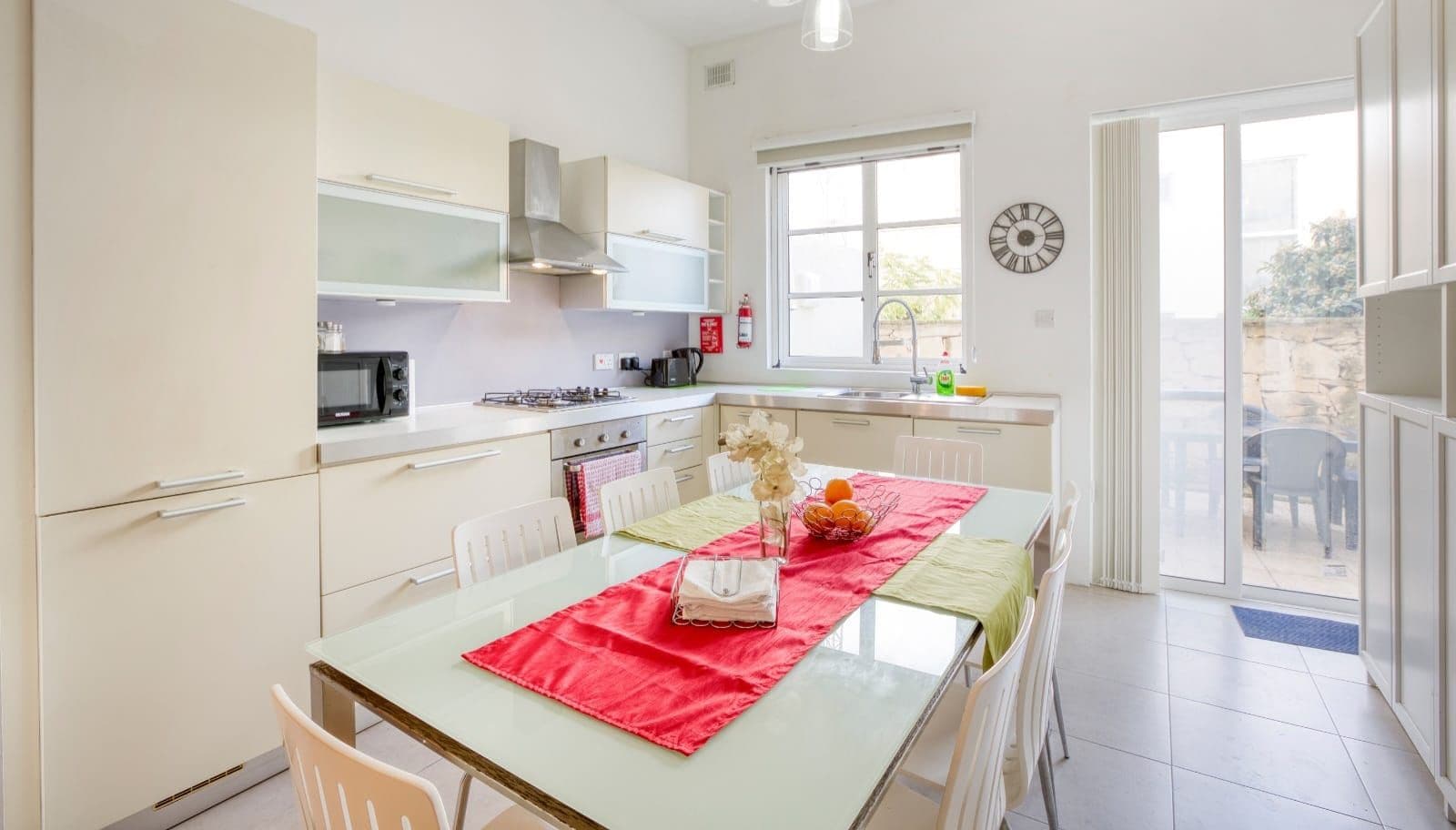 A modern kitchen with white cabinets, a dining table with a red table runner, and a view of the outside through a sliding glass door.