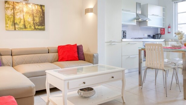 A living room with a beige sofa, a glass coffee table, and a red pillow. There is a kitchen area with white cabinets and a dining table with chairs. - BELAIR (St. Julian's)