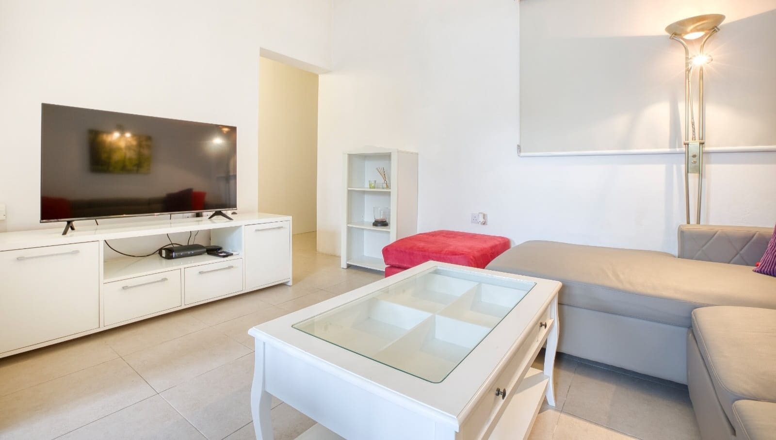 A modern living room with a flat-screen TV, white furniture, a glass-top coffee table, and a red accent pillow.
